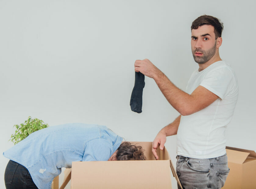 Wife and husband are busy, unpacking the carton boxes. Man is standing. holding black sock, looking at the camera with silly face expression.